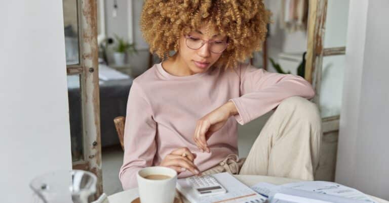 Person with curly hair wearing glasses, sitting at a table with a calculator and documents, focused on managing healthcare finances. The setting appears to be a home office, with a cup of coffee nearby and a relaxed, yet thoughtful atmosphere.