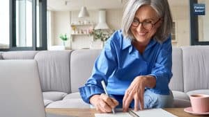 A smiling woman in glasses sitting on a couch and writing notes, likely involved in health research or personal health management.