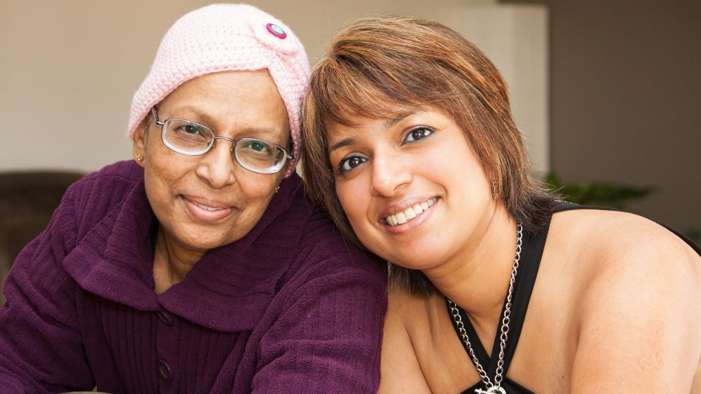 A warm, close-up image of two women, one with a headscarf, smiling at the camera, suggesting a moment of connection in long-distance caregiving.