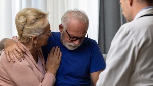 An elderly man, comforted by a woman, receives news from a doctor, capturing a moment of learning about being newly diagnosed and having to manage his diagnosis.