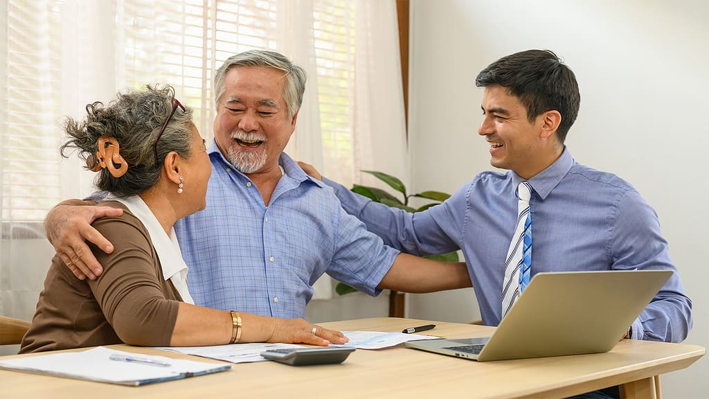 An elderly couple happily discussing options with a representative from a 'home care agency' at a table with a laptop.