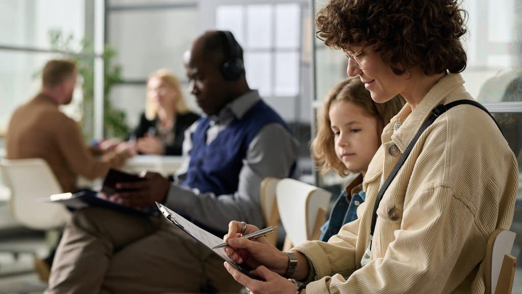 Patients in a waiting room filling out intake forms, with a healthcare provider assisting a young boy in the foreground.