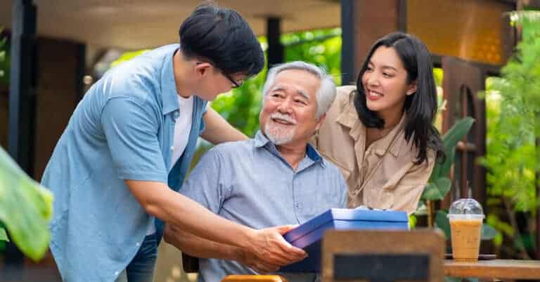 A young healthcare professional provides constructive feedback to an elderly man holding a booklet, as a woman accompanies, in a cheerful clinical setting.