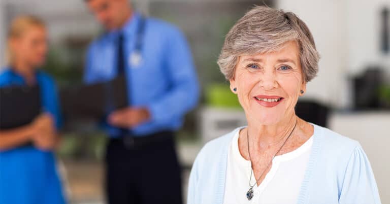 A senior woman smiling at the camera with a healthcare provider in the background, representing the concept of 'Transitional Care.