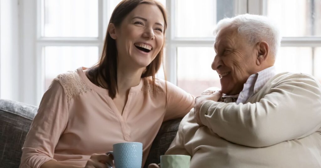 A young woman and an elderly man sit together on a cozy couch, smiling and enjoying a cup of coffee, symbolizing support and companionship in performing activities of daily living, such as social engagement and maintaining personal care routines in a warm, home-like environment.