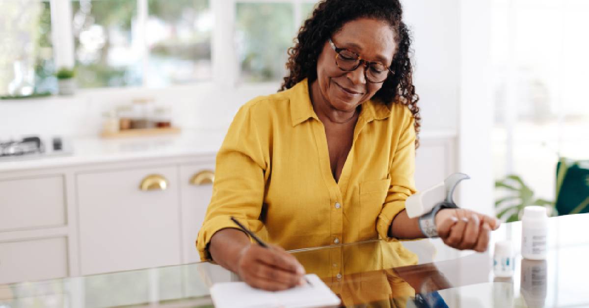 Smiling woman in a yellow shirt sitting at a kitchen table, monitoring her blood pressure and taking notes, illustrating proactive chronic condition management.