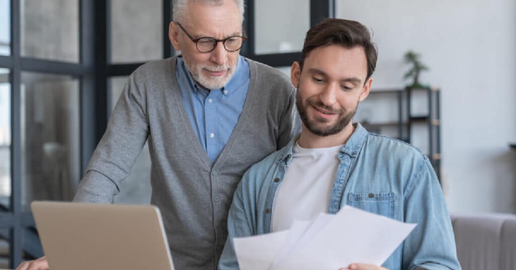 An elderly man and a younger man reviewing documents together in front of a laptop, representing the concept of a Clearinghouse where healthcare data and financial transactions are managed and processed.