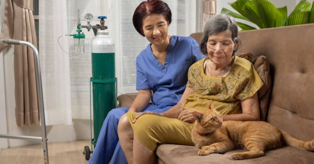 An elderly woman with a nasal cannula for oxygen support sits on a sofa, gently petting a ginger cat beside her. A caregiver in blue scrubs sits next to her, smiling warmly, offering emotional support and companionship. An oxygen tank and walker are visible in the background, indicating a home healthcare setting for managing a chronic disease. The peaceful atmosphere, with soft lighting and indoor plants, highlights compassionate care and the importance of maintaining quality of life for individuals living with chronic illnesses.