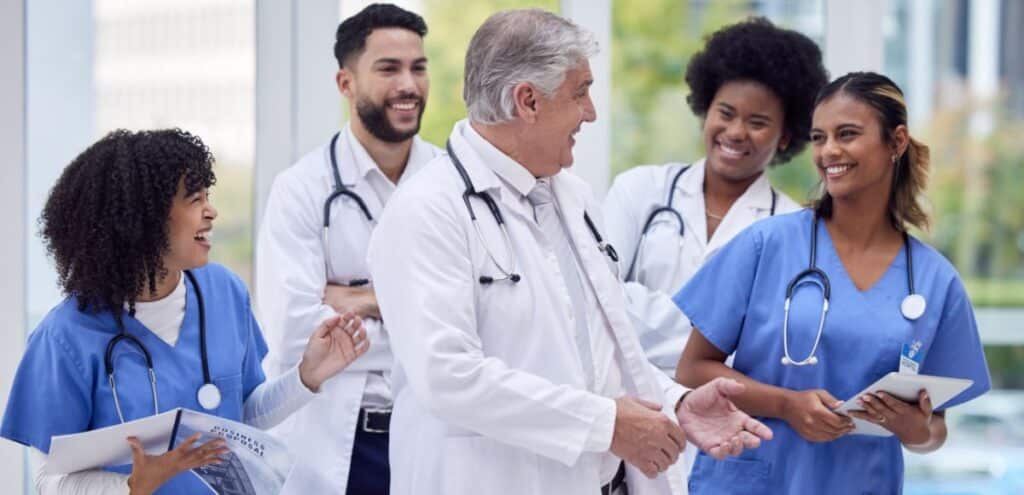 A group of diverse healthcare professionals, including doctors and nurses, engage in a lively discussion in a modern medical facility. The central figure, an older male doctor in a white coat with a stethoscope, gestures confidently as he explains a point, symbolizing leadership in healthcare. The team, dressed in blue scrubs and lab coats, smiles and listens attentively, holding clipboards and tablets. This image highlights collaboration in the healthcare system, which often operates under the fee-for-service model, where individual medical services are billed separately. The bright, professional setting underscores efficiency and dedication to patient care.