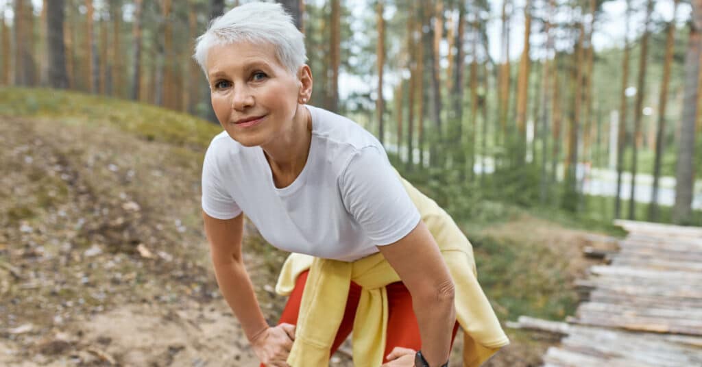 A middle-aged woman in a white t-shirt and red pants is taking a break from her exercise routine in a forest. She is leaning forward with her hands on her knees, smiling, and looking content. This image represents health, showcasing the importance of staying active and enjoying outdoor activities for physical and mental well-being.