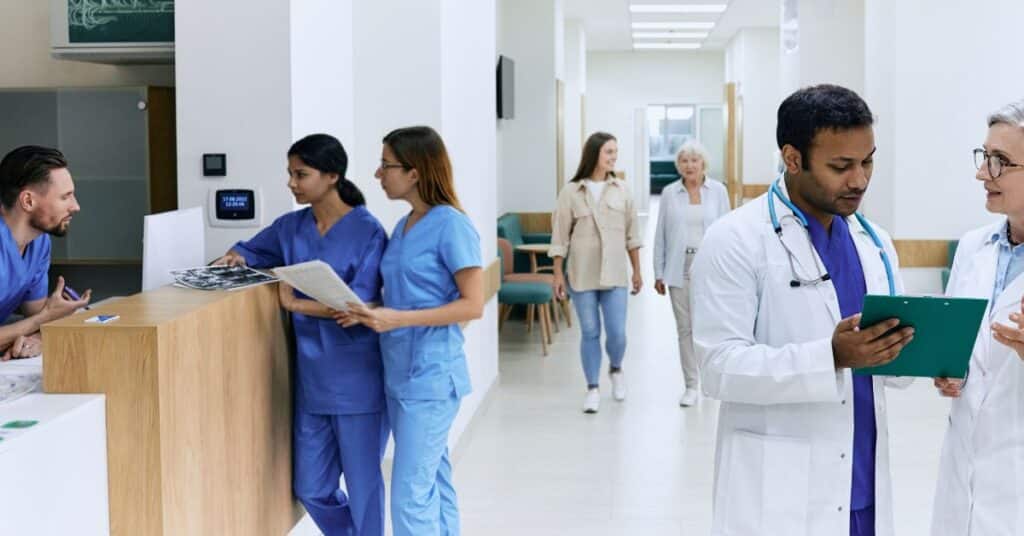 A busy hospital hallway with healthcare professionals and patients interacting. On the left, a group of nurses in blue scrubs are discussing medical images and notes at a reception desk. On the right, two doctors in white coats are engaged in a discussion while reviewing a clipboard. In the background, two patients, including an elderly woman, walk down the hallway. This image highlights the importance of health communication in ensuring effective coordination and collaboration between medical staff, improving patient care, and facilitating a well-organized healthcare environment. The bright, clean setting emphasizes professionalism and clarity in communication.