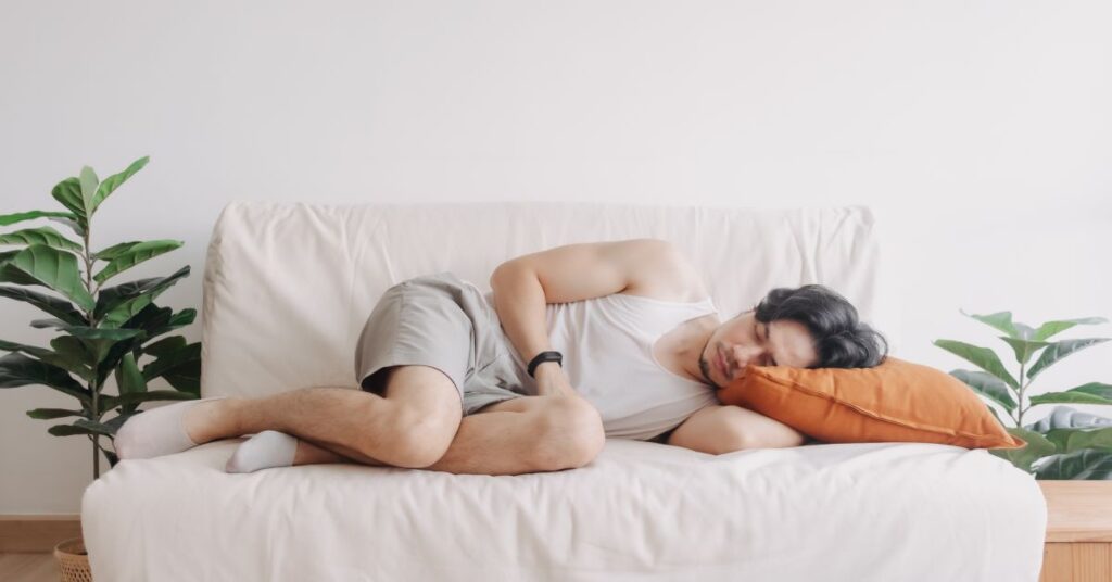 A man is resting on a white sofa in a tranquil home setting, lying on his side with an orange pillow supporting his head. Dressed casually in a white tank top and light gray shorts, he wears a fitness tracker on his wrist, suggesting he is tracking his health status as part of a broader health story. The surrounding space, adorned with green plants, creates a peaceful ambiance that emphasizes the role of rest and mindfulness in personal well-being. This image represents an individual's ongoing health story, focusing on self-care, recovery, and proactive health monitoring in everyday life.