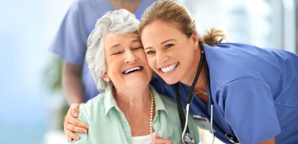 A smiling health care professional wearing scrubs leans affectionately toward an elderly patient, showcasing a warm and compassionate bond. The healthcare provider’s stethoscope is visible, symbolizing medical care and support. The elderly patient, dressed in a light-colored blouse with a pearl necklace, exudes joy and comfort in the presence of the caregiver. The background features a softly lit healthcare setting, emphasizing trust and personalized attention in patient care.