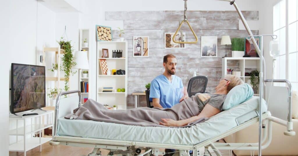 A home health professional in a blue medical uniform provides care to an elderly patient lying in a hospital bed within a bright, homey environment. The room is adorned with personal touches such as plants, framed pictures, and a cozy decor that reflects the comfort of receiving healthcare services at home. Medical equipment like an IV stand and a bed assist handle highlight the specialized nature of home health care, emphasizing the patient\u2019s ability to recover in a familiar and supportive setting. This scene captures the essence of home health, blending professional medical care with the warmth of a personalized home atmosphere.