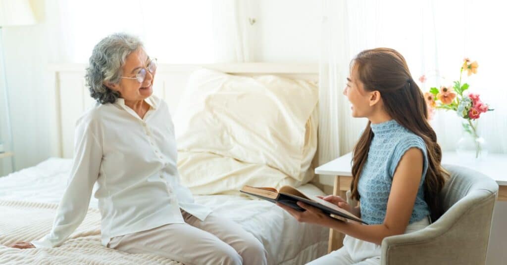 A warm and inviting scene depicting a compassionate caregiver engaged in homecare services with an elderly woman in a cozy bedroom setting. The elderly woman, seated on a neatly made bed with a soft white duvet, smiles warmly at the caregiver, who sits beside her holding an open book. The room exudes a peaceful atmosphere, enhanced by natural light streaming through the windows and colorful flowers placed on a nearby table. This image highlights the personal and comforting nature of homecare, emphasizing support, companionship, and quality of life provided to individuals in their own homes.