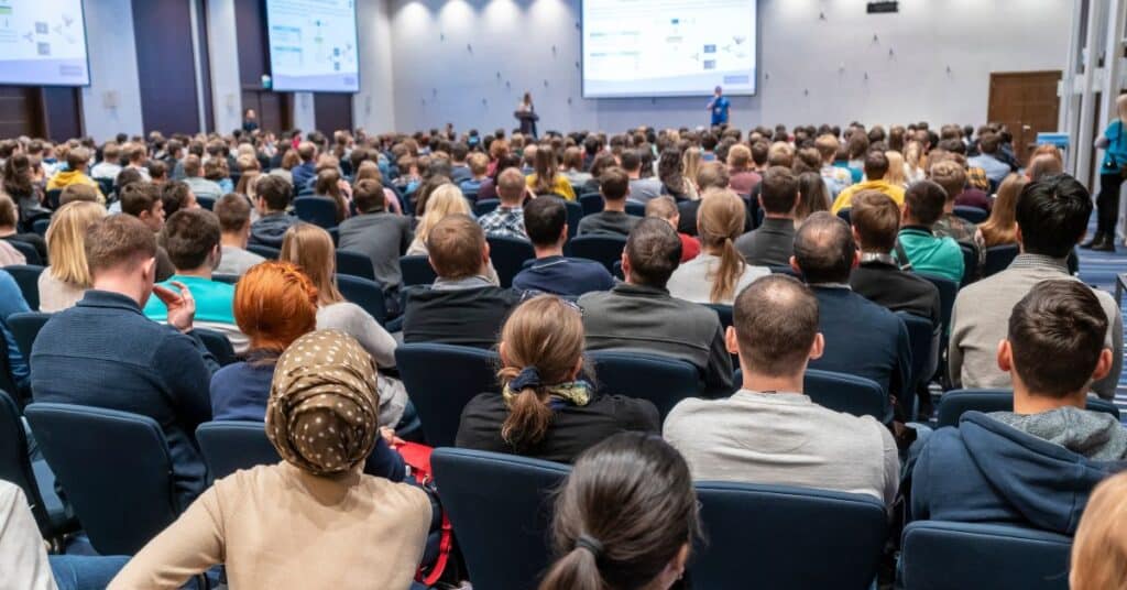 This image shows a large audience attentively listening to a presentation in a conference hall. The setting suggests a professional or educational environment, with diverse attendees seated in rows of chairs, facing a stage where speakers are delivering a talk supported by projected visuals. The scene highlights an atmosphere of engagement and knowledge-sharing, ideal for fostering meaningful learning experiences, collaboration, and professional development.