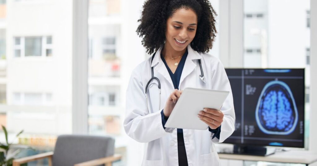 A healthcare professional wearing a white coat and stethoscope stands in a bright medical office, using a digital tablet to review patient information. Behind her, a computer screen displays a brain scan, emphasizing advanced diagnostic tools. Her focused expression and the use of technology highlight the concept of meaningful use, which refers to the effective use of electronic health records (EHRs) to improve patient care, enhance clinical workflows, and ensure better health outcomes. The modern setting, with large windows and natural light, underscores a forward-thinking, technology-driven approach to healthcare.