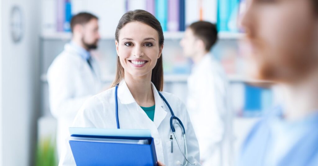 A smiling female medical professional wearing a white coat and stethoscope stands confidently in a brightly lit healthcare setting, holding a blue folder filled with documents. The background features a team of medical practitioners in discussion, symbolizing collaboration and expertise in the medical field. This image highlights professionalism and the role of medical staff in delivering patient-centered care.