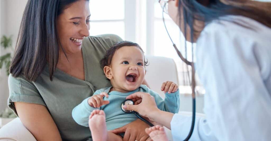 The image captures a joyful moment during a medical care appointment, showing a happy baby sitting on their mother’s lap while a healthcare professional conducts an examination. The doctor uses a stethoscope to listen to the baby’s chest, ensuring the child’s health and well-being. The mother, dressed casually, smiles warmly, creating an atmosphere of trust and comfort in a bright, welcoming clinical environment. This scene highlights the importance of compassionate and attentive medical care in fostering positive healthcare experiences for families.