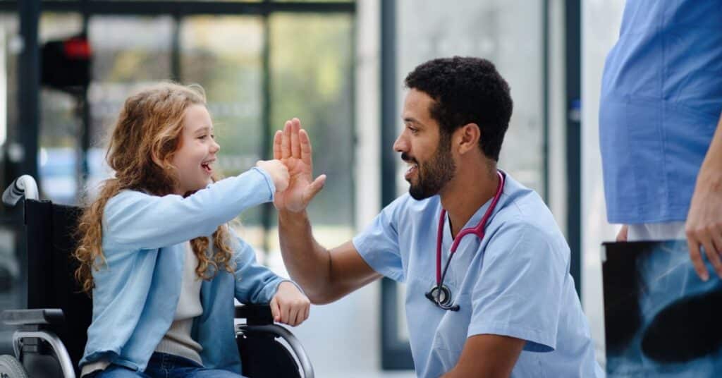 Healthcare professional providing patient care to a young girl in a wheelchair, sharing a high-five moment of encouragement and connection in a brightly lit medical facility, emphasizing trust and compassion in healthcare