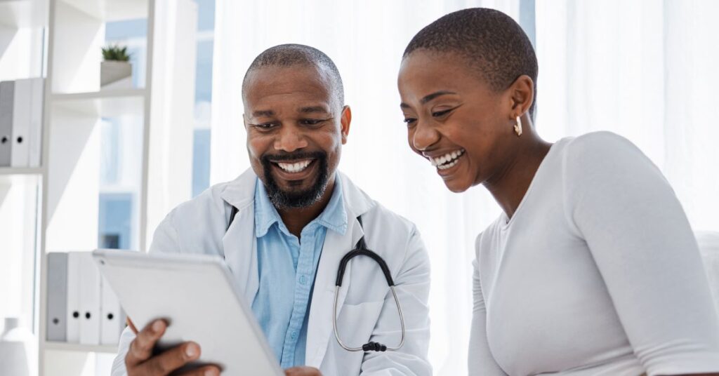 An image showcasing patient engagement in action: A smiling doctor in a white coat and stethoscope holds a tablet, collaborating with an equally joyful patient seated beside them. The interaction reflects mutual trust and open communication, key components of patient engagement. The modern, bright medical office setting highlights the use of technology and patient-centered care to foster informed decision-making and active participation in health management.