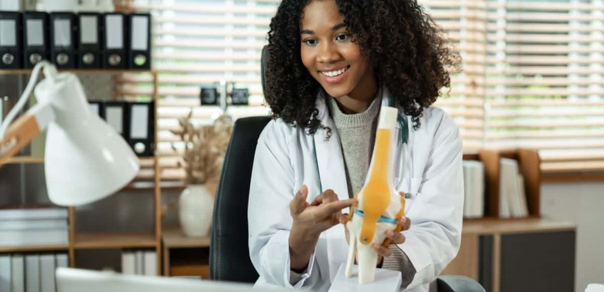 A friendly healthcare professional wearing a white coat and stethoscope sits at her desk, holding a model of a human knee joint and explaining its parts. She gestures toward the model with a warm smile, illustrating the use of medical terminology to describe anatomical structures and treatment options. The well-lit office, with shelves of organized binders and a bright desk lamp, creates a professional yet approachable atmosphere. This image emphasizes the importance of clear communication in healthcare, helping patients understand complex medical concepts.