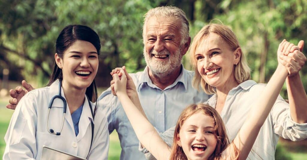 A joyful group consisting of a healthcare professional, an older man, a woman, and a young girl standing outdoors, holding hands, and smiling together. The healthcare provider, wearing a white coat and stethoscope, shares in the positive atmosphere, emphasizing a caring and collaborative connection. The image reflects the concept of Relationship-Centered Care, where healthcare focuses not just on the individual patient, but on fostering meaningful partnerships between providers, patients, and their families to achieve better health outcomes and a more compassionate care experience. The vibrant greenery in the background adds a sense of warmth and well-being.