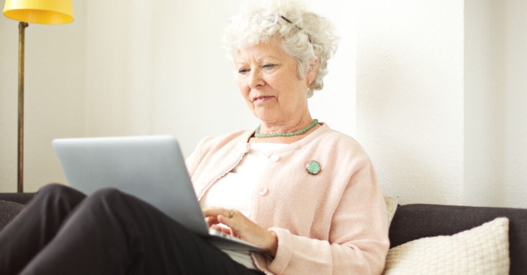 An older woman with curly white hair, wearing a soft pink cardigan and a green necklace, sits comfortably on a couch, using a laptop. She appears focused and engaged, symbolizing active participation in her healthcare from the comfort of her home. A warm lamp and pillow in the background add to the cozy, relaxed setting. This image represents Remote Patient Monitoring, highlighting how patients can use digital technology to share health information with their healthcare providers in real-time, enabling continuous care and reducing the need for in-person visits.