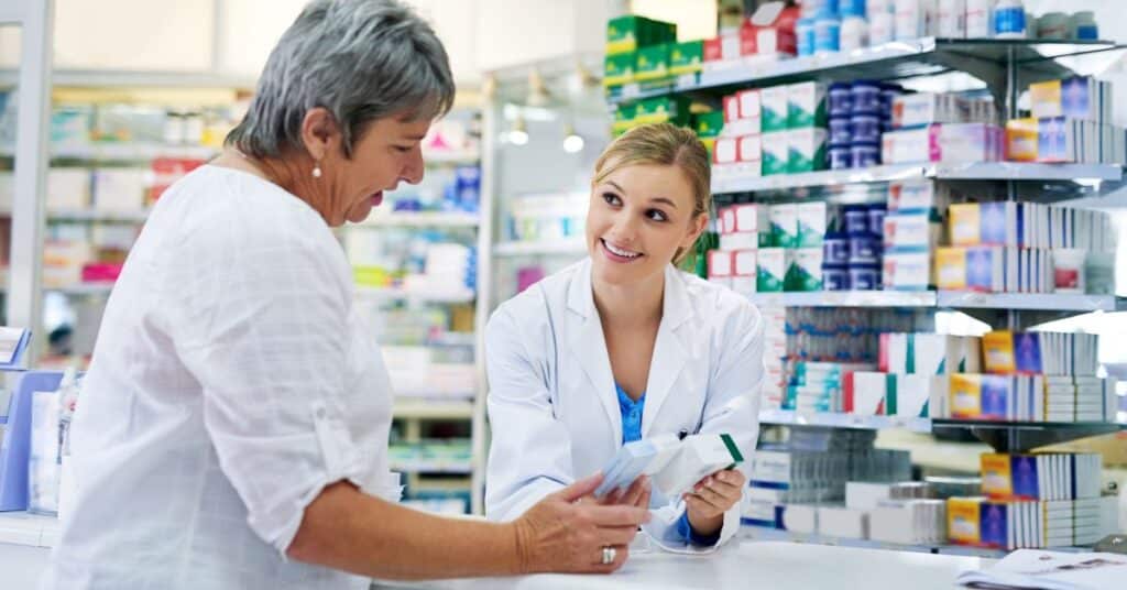 The image depicts a retail health clinic setting where a friendly healthcare professional, wearing a white lab coat, is attentively assisting an older woman. The professional is holding a boxed medication, explaining its use, while the woman listens with interest. Behind them, shelves are stocked with an array of over-the-counter medicines and healthcare products, emphasizing the accessible and convenient nature of retail health clinics. This environment highlights the role of retail health clinics in providing immediate care, routine health services, and personalized consultations within a retail pharmacy setting.