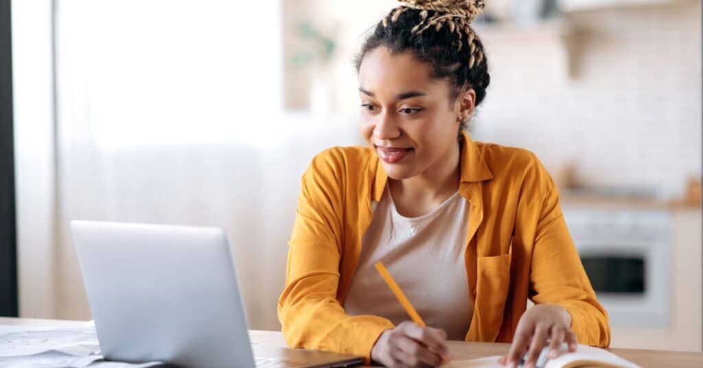 The image depicts a young woman engaging in rote learning at home. She sits at a table with a laptop open in front of her, holding a pencil and writing in a notebook. Her focused expression and tidy workspace suggest she is memorizing information through repetition. The bright, natural light from the window highlights the casual yet productive atmosphere, emphasizing the practical application of rote learning for studying or mastering foundational knowledge.