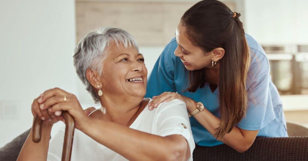 The image captures a warm and supportive interaction between an elderly woman and a caregiver in a home or care facility setting. The elderly woman, seated and holding a wooden cane, is smiling brightly, exuding a sense of joy and comfort. Beside her, the caregiver, dressed in professional scrubs, leans in with a gentle smile, placing her hands reassuringly on the woman's shoulders. The natural lighting and casual yet intimate environment suggest trust and companionship, reflecting the importance of emotional connection and personalized care in caregiving relationships.