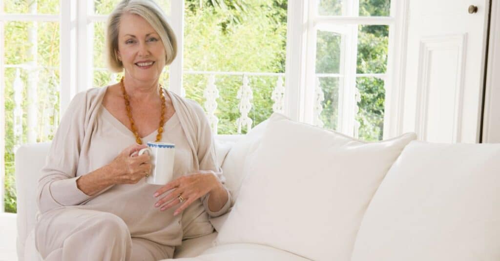 A serene older woman sitting comfortably on a white sofa in a bright, airy living room, holding a cup of tea and smiling softly. Dressed in light, cozy attire, she exudes a sense of calm and relaxation. The large windows behind her offer a view of lush greenery, adding to the peaceful ambiance. This image represents self-care, illustrating the importance of taking time to relax, recharge, and nurture one's well-being in a comfortable environment.