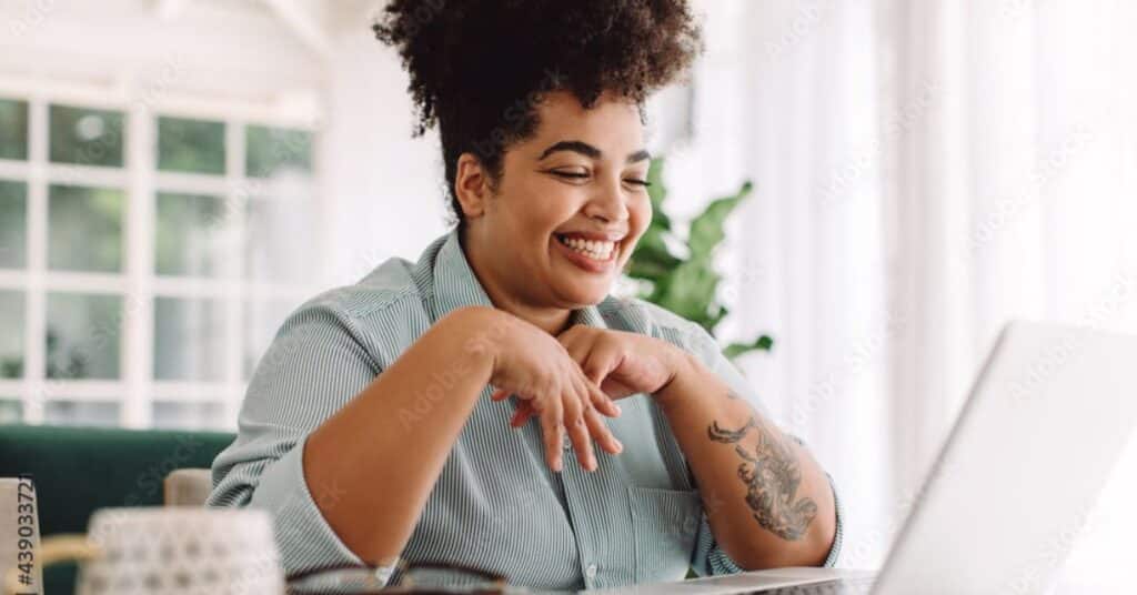 A smiling woman with curly hair, wearing a striped shirt, sits at a desk in a bright, cozy room, engaging with her laptop. Her body language shows excitement and confidence as she interacts with the screen, symbolizing proactive learning and engagement. The calm setting, with natural light streaming through large windows and a plant in the background, reflects a supportive environment for self-health management. This image represents the concept of individuals taking charge of their healthcare by using digital tools and resources to learn, track, and manage their health effectively.