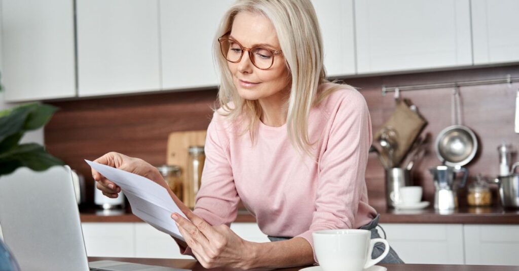 A mature woman with long blonde hair and glasses is standing in a modern kitchen, carefully reviewing a document while working on her laptop. A cup of coffee sits on the counter beside her, creating a calm and focused atmosphere. Her attentive expression suggests she is taking an active role in organizing and understanding her healthcare information. This image illustrates the concept of self-managing care, where individuals take responsibility for tracking their medical records, understanding treatment plans, and making informed healthcare decisions in their daily lives.