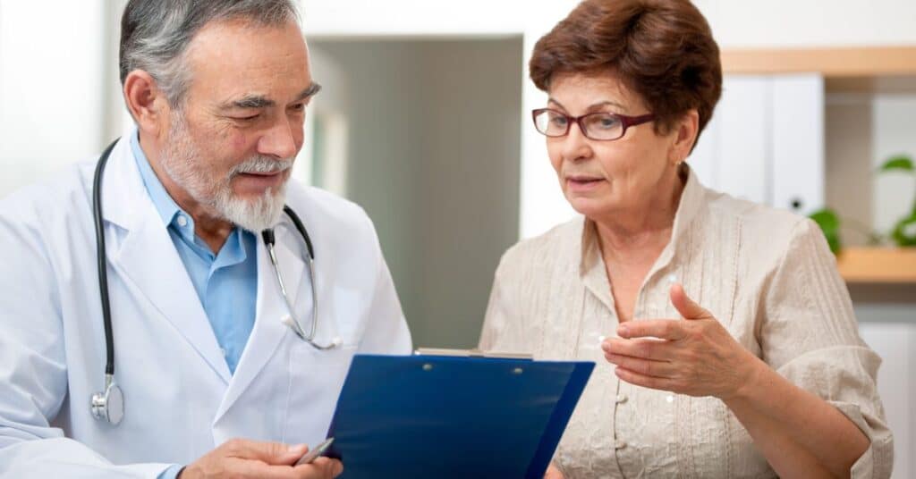 An older woman consulting with her doctor, who is holding a clipboard and explaining medical details. This image illustrates the concept of being 'under a physician's care,' emphasizing the importance of professional medical guidance and collaborative decision-making in managing health conditions.