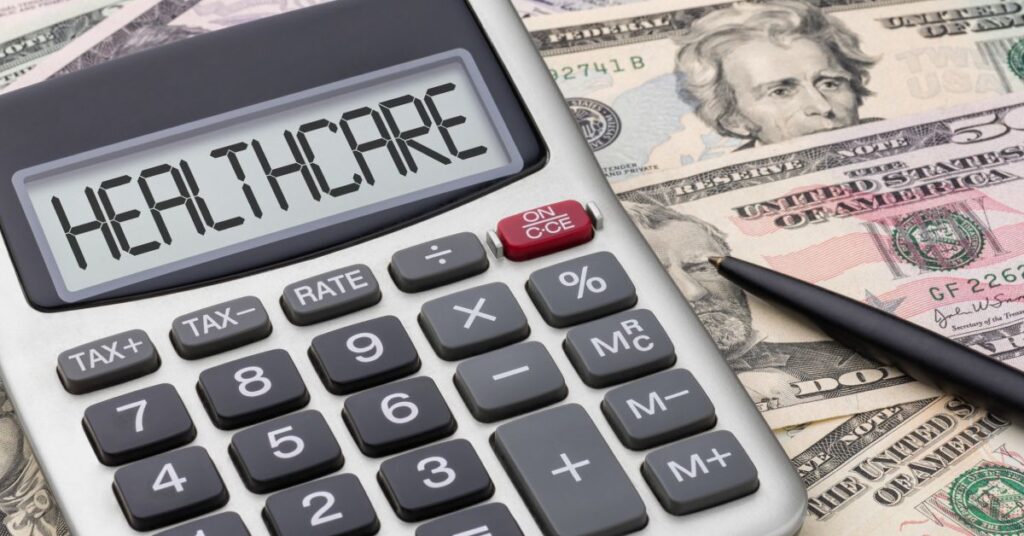 The image features a close-up of a calculator displaying the word “HEALTHCARE” on its screen, symbolizing the calculation and analysis of healthcare costs. The calculator rests on a pile of U.S. currency, including $20 bills, emphasizing the financial considerations associated with medical expenses. A black pen lies beside the calculator, suggesting the need for planning, budgeting, and decision-making in managing healthcare expenses. This visual highlights the importance of understanding healthcare costs and the tools required to navigate the financial aspects of medical care effectively.