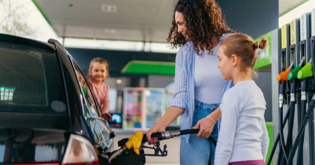 A mother pumping gas into her car while her children watch, symbolizing the concept of indirect costs in healthcare such as transportation expenses.