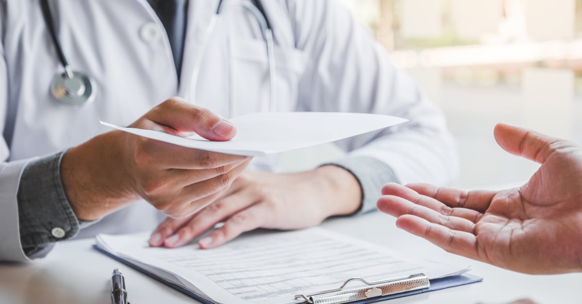 A doctor handing a patient a prescription in a clinical setting, with medical forms and a clipboard on the table. The focus is on the interaction between the doctor and patient, highlighting the importance of understanding medical abbreviations for clear communication and effective healthcare management.