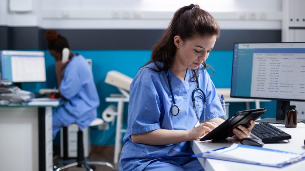 A nurse in blue scrubs reviews patient information on a digital tablet in a modern clinical office, while another healthcare professional speaks on the phone in the background. Computer monitors displaying patient data surround the workspace, illustrating the organized and technology-driven environment often associated with pay-for-performance healthcare models, where quality and efficiency in care delivery are prioritized to achieve better patient outcomes.