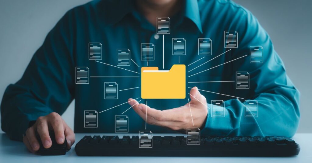 The image depicts a professional sitting at a desk, symbolizing the concept of organizing and managing documents. At the center is a prominent yellow folder icon surrounded by multiple connected document icons, representing a centralized document management system. The individual's hands are slightly raised, as if presenting or supporting the digital organization process, while the keyboard beneath suggests active engagement in creating or maintaining an electronic filing system. The visual highlights the importance of efficiently organizing and accessing documents for streamlined workflows and productivity in various professional settings.