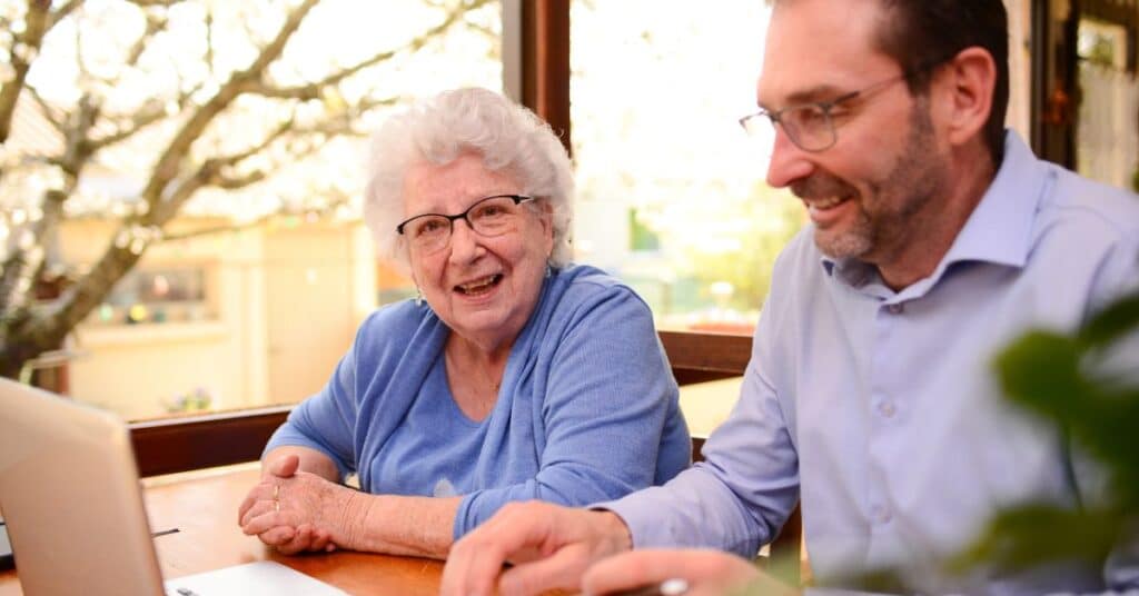 The image features an elderly woman sitting at a table alongside a middle-aged man, engaged in what appears to be a discussion about important documents on a laptop. This setting reflects a Power of Attorney scenario, where the elderly woman, likely the principal, is working with her trusted agent or attorney-in-fact to manage financial, legal, or healthcare decisions. Their collaborative interaction, framed by a cozy, naturally lit environment, emphasizes the importance of trust, communication, and careful planning in utilizing a Power of Attorney to ensure the principal’s needs and preferences are upheld.