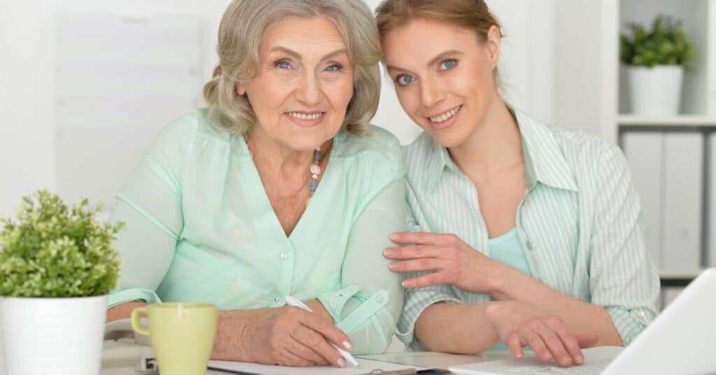 An elderly woman and her primary caregiver smile together at a desk. The elderly woman, with gray hair and a warm expression, is writing in a notebook while her primary caregiver, a young blonde woman, sits beside her with one hand on her shoulder, looking at a laptop. They are both dressed in light green attire, and the setting appears to be a bright, comfortable home or office space with plants and shelves in the background.