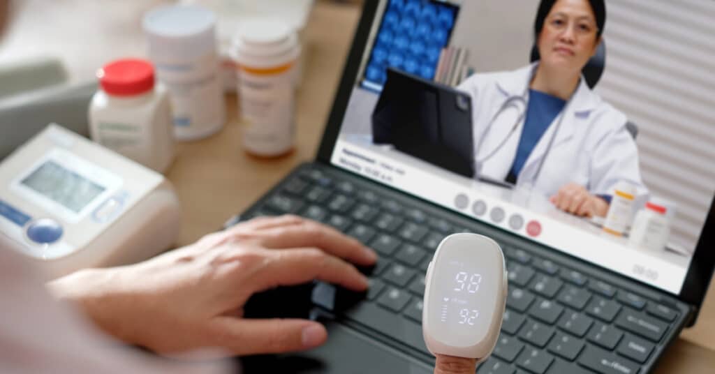 A patient is interacting with a healthcare provider through a video call on a laptop in a virtual setting. The patient is using a pulse oximeter, with various medications and a blood pressure monitor visible on the desk. This scene highlights the use of a virtual setting for remote medical consultations and health monitoring.