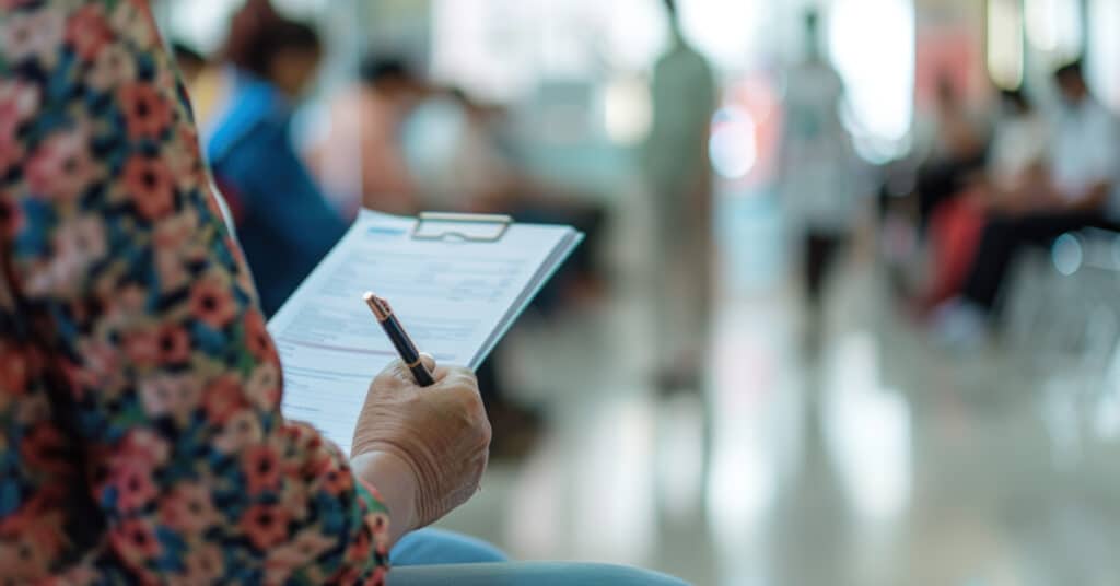 A person holding a clipboard with a health history document in a busy medical waiting room. The clipboard contains detailed sections for personal information, medical history, medications, allergies, immunizations, and recent doctor visits, illustrating the concept of what is health history.