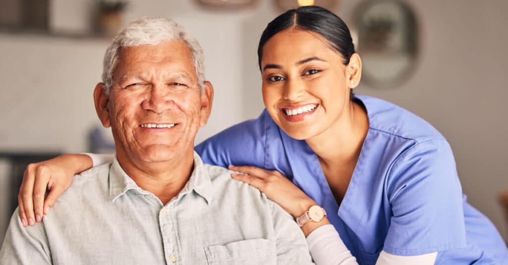 A smiling elderly man and a young female caregiver in a home care setting. The caregiver, dressed in blue scrubs, is standing behind the man, with her arms gently resting on his shoulders. Both are smiling warmly, reflecting the positive and supportive environment of home care.