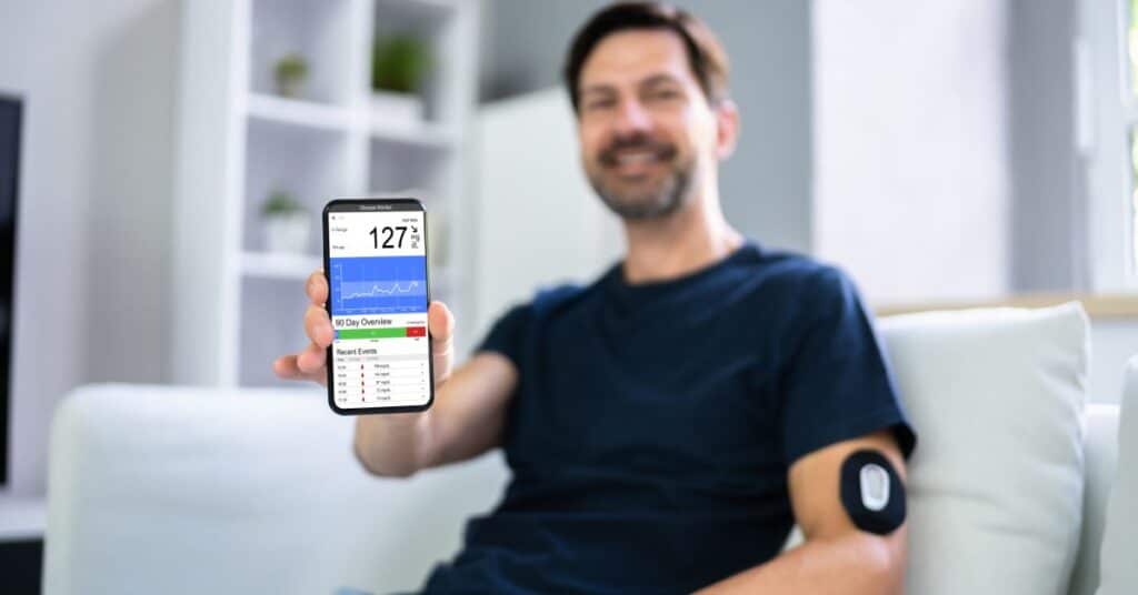 The image shows a man sitting comfortably on a couch, holding up his smartphone displaying remote patient monitoring app with glucose level readings. The screen indicates a glucose level of 127 mg/dL with a 90-day trend chart and recent data points below it. The man is also wearing a wearable glucose monitoring device on his arm, which is seamlessly transmitting real-time health data to the app. This setting highlights the convenience and effectiveness of remote patient monitoring technology, empowering individuals to actively track and manage their health metrics from home.