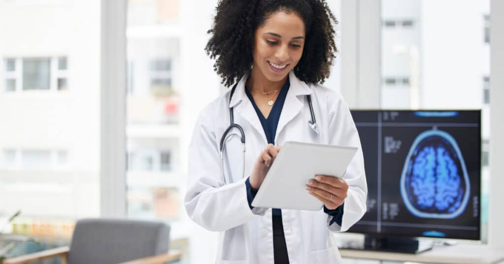 A healthcare professional in a modern office, smiling and reviewing medical data on a tablet. Behind her, a computer screen displays medical images. The scene illustrates the store-and-forward telecommunication method in healthcare, where medical data is collected and securely transmitted for later assessment by a healthcare provider.