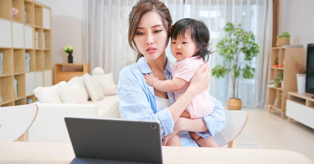 A woman at home, sitting at a table and holding a young child, engaging in a video call on a tablet. The cozy, well-lit living room emphasizes the concept of Synchronous Telecommunication, showcasing real-time communication and immediate interaction between the woman and a remote healthcare provider.