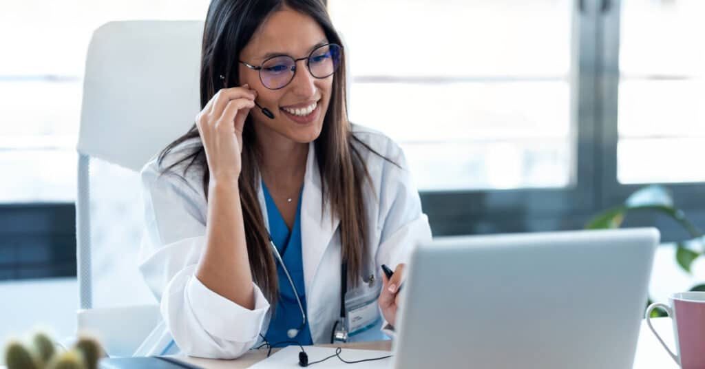 A healthcare provider wearing a headset, sitting at a desk and engaging in a video call on a laptop. The setting is a modern office, and the provider is smiling, emphasizing the use of telecommunication in healthcare to facilitate real-time consultations and communication with patients.