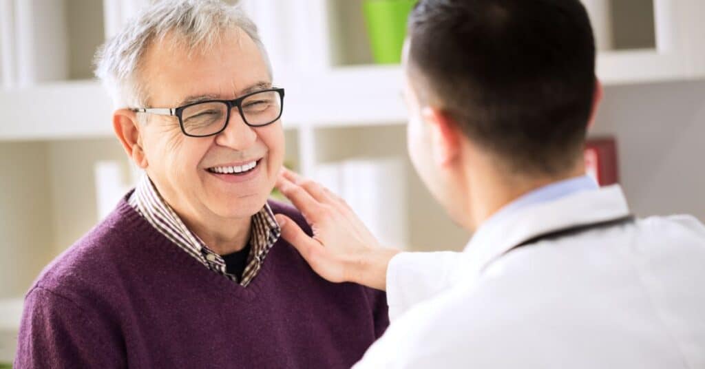 An elderly man with glasses, wearing a purple sweater, smiles warmly while sitting in a doctor's office. A healthcare provider, dressed in a white coat, places a reassuring hand on the man's shoulder, illustrating trust and care. This image reflects the definition of a patient by Patient Better: an individual receiving medical care, treatment, or consultation for an illness, injury, or health condition in a professional healthcare setting.